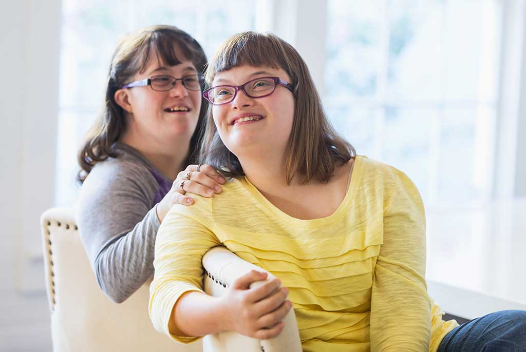 Two sisters with down syndrome sitting on chairs at home by windows.