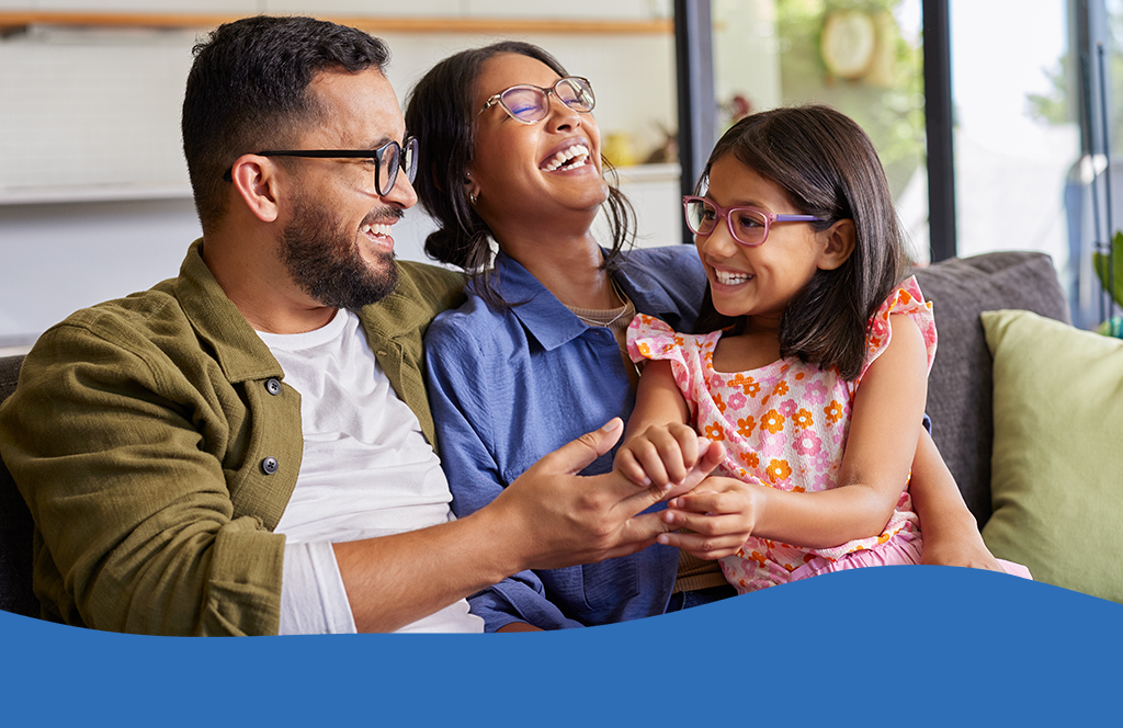 Cheerful middle eastern family with eyeglasses of three having fun together at home. Mixed race parents talking with their little girl in living room. Cheerful family with specs embracing and playing with beautiful indian daughter girl in arms.